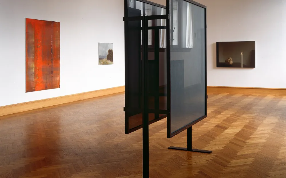 Ein heller Ausstellungsraum mit Parkettboden: an den Wänden hängen Bilder, in der Mitte stehen zwei freistehende Rahmen auf schmalen Füßen.